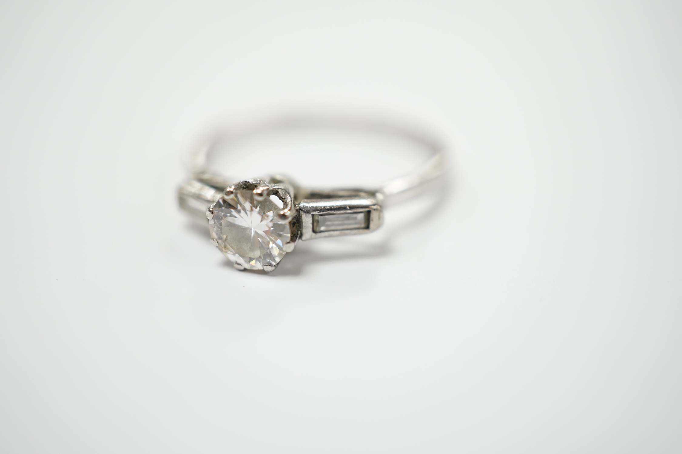 A white metal and single stone diamond set ring, with two stone baguette cut diamond set shoulders, size O, gross weight 2.3 grams, stone diameter 5.2mm.
